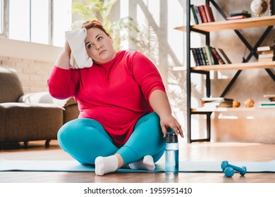Tired exhausted young caucasian fat woman sitting and relaxing after hard training at home for fat burning and losing weight. Chubby plump woman doing fitness exercises indoors
