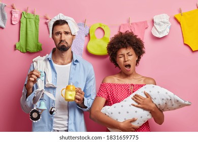 Tired exhausted young mother poses with baby on hand has sleepless nights while takes care of her newborn daughter. Displeased father holds feeding bottle and mobile helps wife with child upbringing