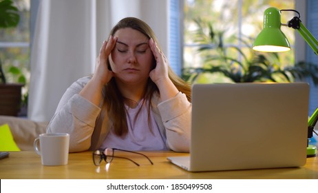 Tired Fat Woman Suffering Eyestrain Working On Laptop At Home. Portrait Of Obese Young Female Studying At Home Having Migraine And Massaging Temples Sitting At Table With Computer