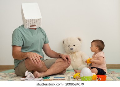 Tired Father With A Toy Box On Head While Is Playing With His Baby Girl On A Play Mat. 