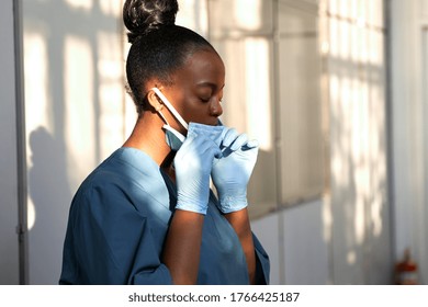 Tired female african scrub nurse wear blue uniform gloves taking off face mask to breathe in hospital. Exhausted black doctor feels stress relief concept, burnout fatigue at difficult work in hallway.