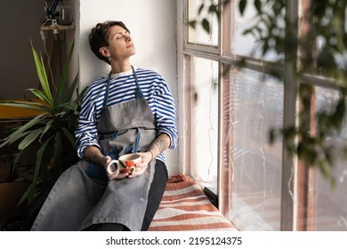 Tired female artisan with cup of tea in hands on windowsill, resting in ceramics studio, dreaming to open her own pottery studio. Peaceful female ceramist taking break during workday in workshop