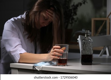 Tired Female Doctor Sitting At The Table And Hold Glass With Whiskey Or Cognac At Home After Hard Work, Depressed Female Drinking Strong Alcohol Suffering From Coronavirus Problems.