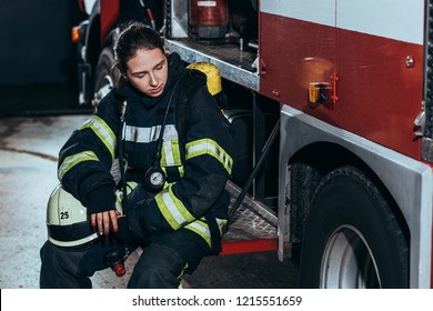 Tired Female Firefighter In Uniform With Helmet Sitting On Truck At Fire Station