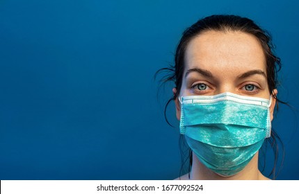 Tired Female Nurse, Wearing A Turquoise Surgical Mask, Exhausted After A Difficult Day In The Hospital Helping  COVID-19, Corona Virus, Patients. Standing Against A Blue Wall With Slogan, Copy Space.