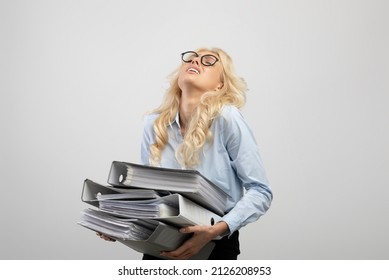 Tired female office worker holding folders of documents, overloaded with paperwork, standing on light grey studio background. Young company employee feeling stressed - Powered by Shutterstock