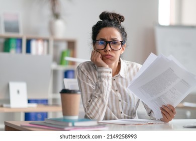 Tired female office worker looking at camera and holding documents, sitting at desk in office. Unhappy woman feeling stressed because of lot of paperwork - Shutterstock ID 2149258993