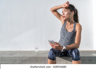 Tired fitness woman sweating taking a break listening to music on phone after difficult training. Exhausted Asian runner dehydrated feeling exhaustion and dehydration from working out at gym.