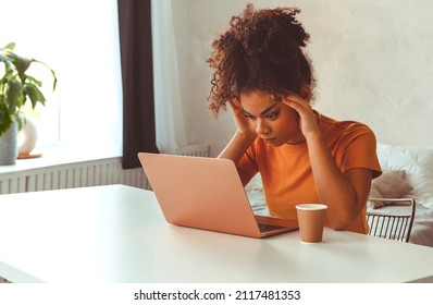 Tired flustrated African descent young girl sitting at desk in front of laptop while irritably looking at computer screen with hands holding her head at temples. Emotional side of freelancing - Shutterstock ID 2117481353