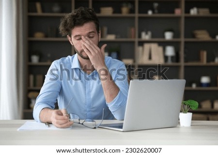 Tired freelance business man sitting at laptop, taking glasses off, touching closed eyes, rubbing eyelids, nose, face, feeling exhausted, overworked, having vision problems, headache
