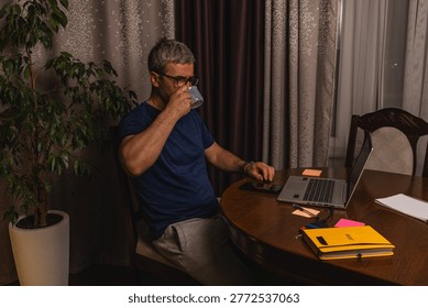 A tired freelancer drinking coffee while working late at night on a laptop in a home office, showing burnout, stress and remote work lifestyle