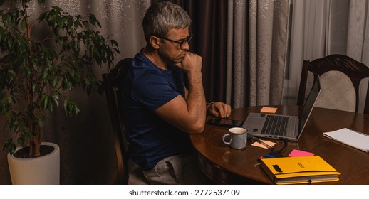 A tired freelancer drinking coffee while working late at night on a laptop in a home office, showing burnout, stress and remote work lifestyle