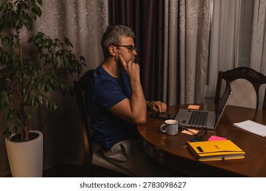 A tired freelancer drinking coffee while working late at night on a laptop in a home office, showing burnout, stress and remote work lifestyle