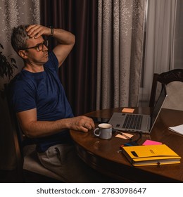 A tired freelancer drinking coffee while working late at night on a laptop in a home office, showing burnout, stress and remote work lifestyle