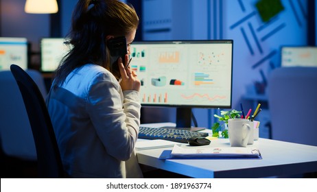 Tired freelancer talking at smartphone with colleague working overtime sitting at desk in business office late at night resolving financial problems. Busy employee using modern technology network