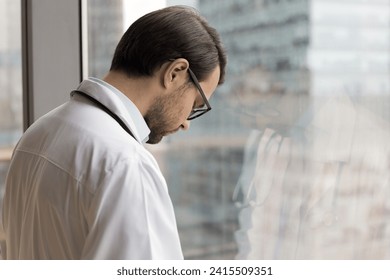 Tired frustrated young doctor man standing at window with closed eyes and head bended down, thinking on problems, practitioner mistake, failure, loss of patient, feeling despair, stress