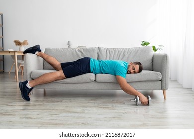Tired funny millennial muscular european man lies on sofa with dumbbell after training in living room interior. Sports at home, workout and new normal and exercises, active lifestyle, rest and relax