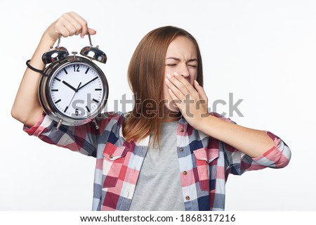 Tired girl holding big alarm clock yawning, isolated over white background. Time concept
