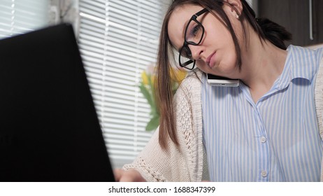 Tired Girl At Kitchen With Glasses Holds Uncomfortable Phone Talks To Someone While Simultaneously Typing Something With Both Hands Into Laptop. Freelancer Girl At Work Business Conversation Concept