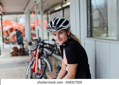 Tired But Happy, Exhausted After Long Training Ride Female Rider Smiles Into Camera In Her Helmet And Lycra Cycling Kit, With Professional Road Bikes In Distance