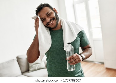 Tired But Happy. Young Man Smiling At Camera, Holding Water Bottle And Wiping Sweat With Towel While Resting After Training Workout At Home. Sport, Healthy Lifestyle. Horizontal Shot