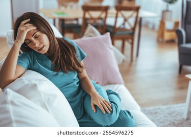 A tired healthcare professional in scrubs rests on a couch, reflecting fatigue and stress after a demanding shift. The cozy home setting contrasts with her exhaustion, illustrating the challenges face - Powered by Shutterstock