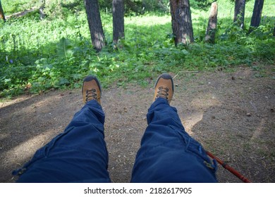 Tired hiker on rest, legs dressed in blue trousers and brown hiking boots with trekking pole nearby. Feet of adult wearing boots to travel walking in green forest. Travel and hiking tourism concept.