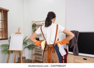 Tired household clean up, housekeeper asian young woman stand and look many clothes on television, cleaning in living room at home. Mess maid or housewife organize dirty and untidy