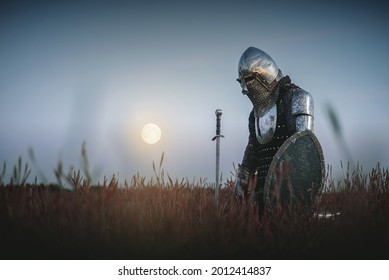The tired knight in the plate armor kneels among the battlefield with a sword in the moon light. Lost battle. Defeat concept.