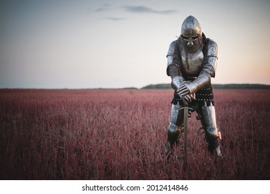 The tired knight in the plate armor stands among the battlefield with a sword. Lost battle. Defeat concept.
