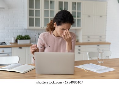 Tired latin female sit at home workplace distracted from laptop hold glasses in hand rub nose bridge. Exhausted young woman freelancer overloaded by computer work feel eye pain strain blurred vision