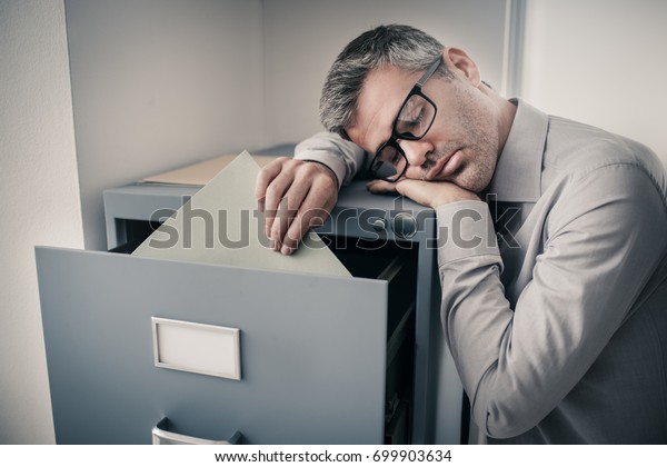 Tired lazy office worker leaning on a\
filing cabinet and sleeping, he is falling asleep standing up;\
stress, unproductivity and sleep disorders\
concept