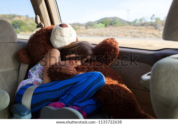 A tired little\
girl rides, curled up in her booster seat atop of her big, stuffed\
bear, in a minivan across the Southwest desert.  She looks bored\
and sad on her long roadtrip.