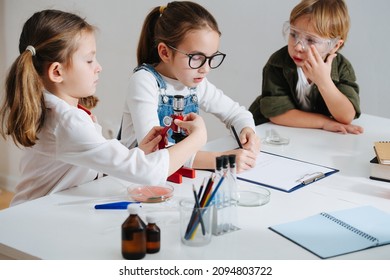 Tired Little Kids Doing Home Science Project, Girl Is Documenting The Process. All Wearing Glasses, Sitting Behind The Table. Chemical Stuff On The Table.