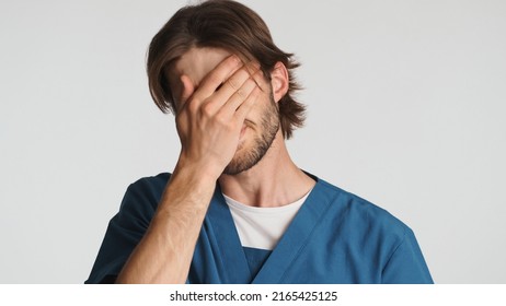 Tired male doctor keeping hand on face something went wrong at work in hospital. Young intern dressed in uniform looking exhausted over white background