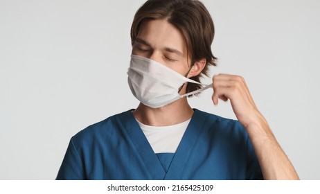 Tired male doctor taking of the medical mask feeling sleepy after hard day in hospital. Young intern dressed in uniform in studio. Overworked expression