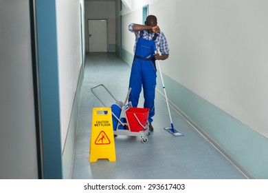Tired Male Janitor With Cleaning Equipment And Wet Floor Sign