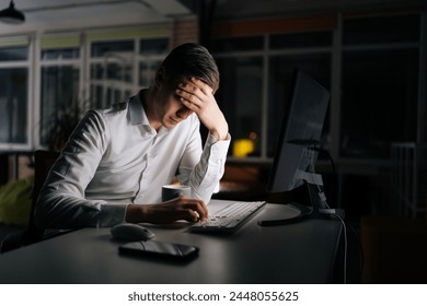 Tired male office worker working late at night, using desktop computer and touching forehead. Exhausted businessman working on laptop computer until night. Concept of stressful life and deadline.