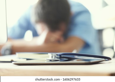 Tired Man Doctor Sleeps Lying On His Hands While Sitting At A Computer Desk. Doctor Works On A Computer In The Clinic After A Night Shift.