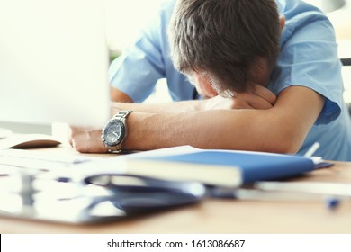 Tired Man Doctor Sleeps Lying On His Hands While Sitting At A Computer Desk. Doctor Works On A Computer In The Clinic After A Night Shift.