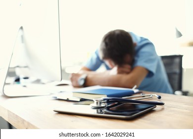 Tired Man Doctor Sleeps Lying On His Hands While Sitting At A Computer Desk. Doctor Works On A Computer In The Clinic After A Night Shift.