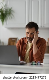 Tired Man Looking At Blurred Laptop In Kitchen And Suffering From Headache