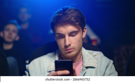 Tired Man Looking Cellphone Screen During Film In Cinema. Portrait Of Bored Guy Surfing Internet On Smartphone In Dark Hall. Closeup Busy Male Person Using Mobile Phone In Movie Theater.