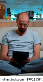 Tired Man Sitting Alone On Couch While Browsing On Internet Using Tablet Computer With Modern Technology Wireless. Caucasian Male In Pajamas Relaxing In Front Of Televion Late At Night In Kitchen