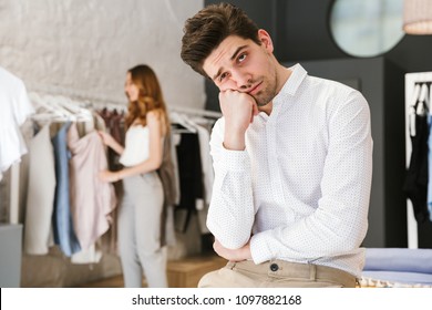 Tired man sitting and looking at camera while his girlfriend shopping for clothes on a background - Powered by Shutterstock