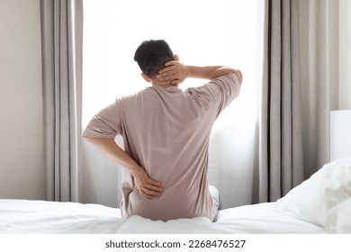 Tired man suffering from back pain after sleeping in bedroom, waking up in morning, sitting on bed and touching lower back and neck, experiencing body aches, wearing pajamas, copy space, back view - Powered by Shutterstock