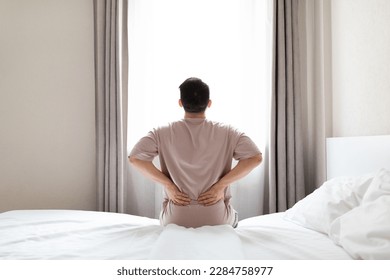 Tired man suffering from back pain after sleeping in bedroom, waking up in morning, sitting on bed and touching his lower back, experiencing muscle strain, wearing pajamas, copy space, back view - Powered by Shutterstock