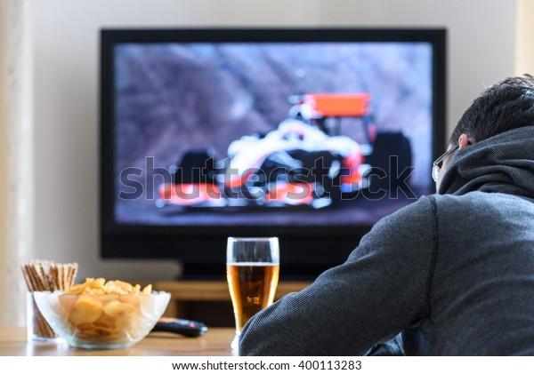 Tired man watching TV (formula one\
race) in living room with alcohol and snacks - stock\
photo