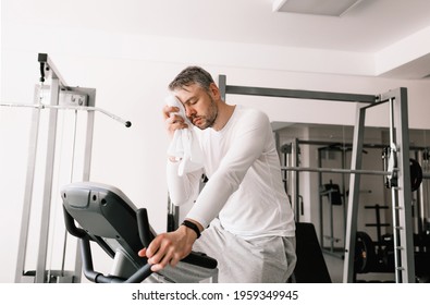 A Tired Man Wipes Sweat From His Face With A Towel During An Exercise On An Exercise Bike. Cardio Workout