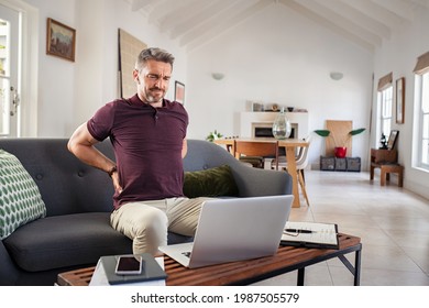 Tired mature businessman stretching back while working from home. Business man suffering from back pain while working on laptop. Stressed man taking a break after long hours at work sitting on couch.